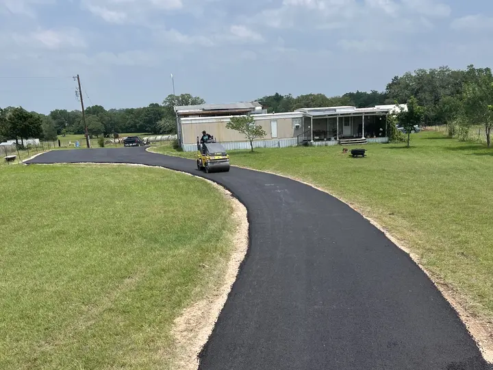 New asphalt driveway curving through green pasture while a roller finishes near the house