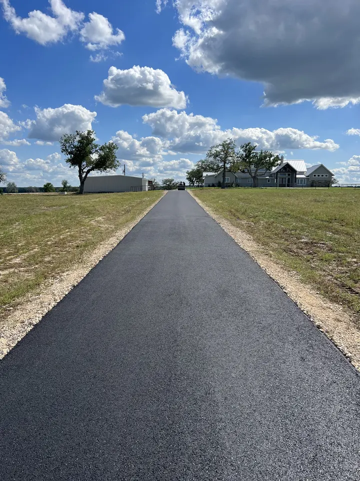 New black asphalt driveway running straight through pasture toward a house under a big Texas sky